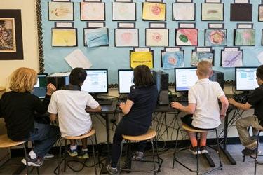 Five students work at computers side by side in a classroom